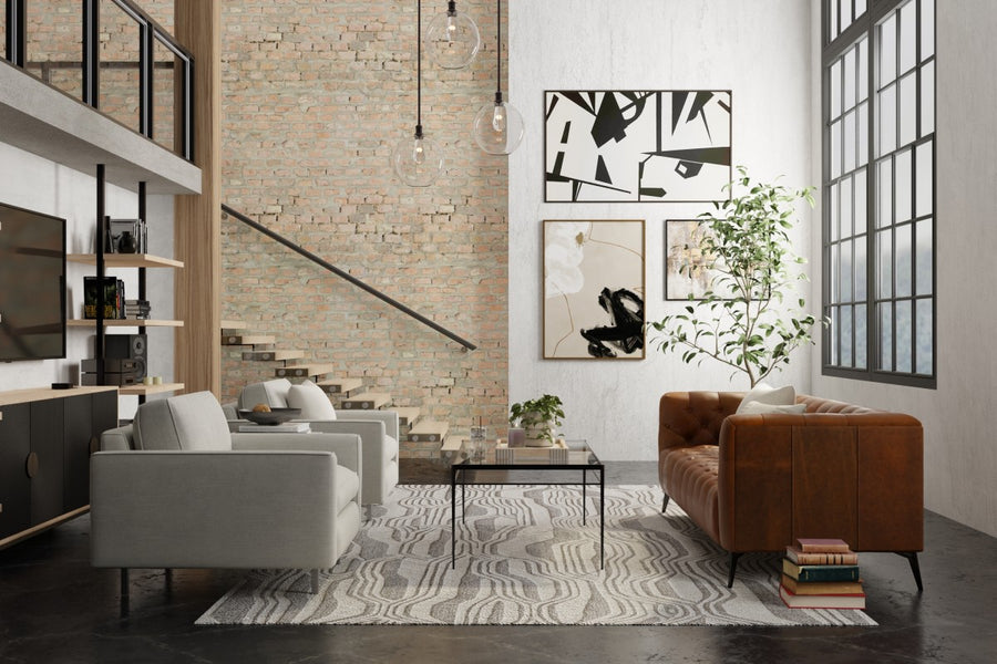 Modern living room with a brown leather sofa, two gray armchairs, a coffee table on a patterned rug, exposed brick wall, abstract art, large windows, and a staircase with a black metal railing.
