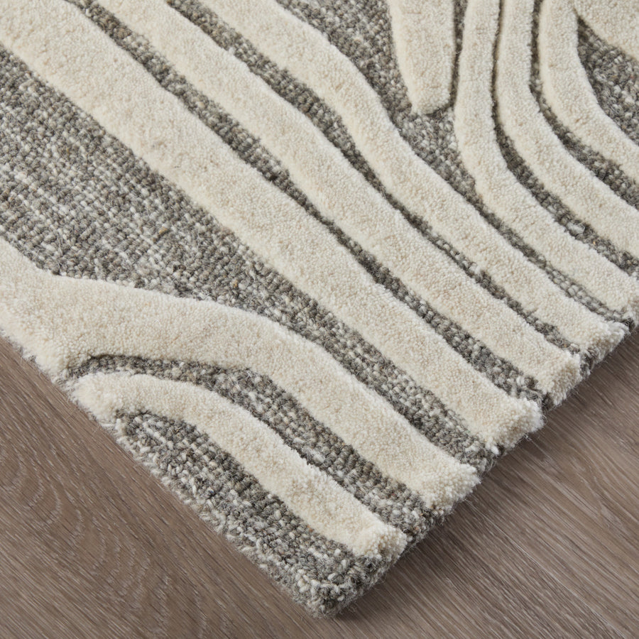 A close-up of a textured area rug with raised, cream-colored lines forming an abstract pattern on a gray background, placed on a light wood floor.