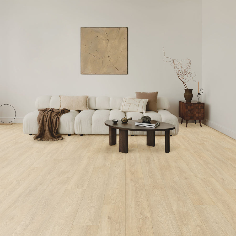 A modern living room with a low, white, tufted sofa, neutral cushions, a brown throw, a round dark coffee table with decor items, a wooden side cabinet, and abstract art on a light beige wall. Light wood floor throughout.