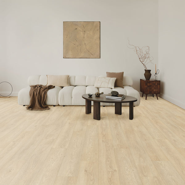 A modern living room with a low, white, tufted sofa, neutral cushions, a brown throw, a round dark coffee table with decor items, a wooden side cabinet, and abstract art on a light beige wall. Light wood floor throughout.