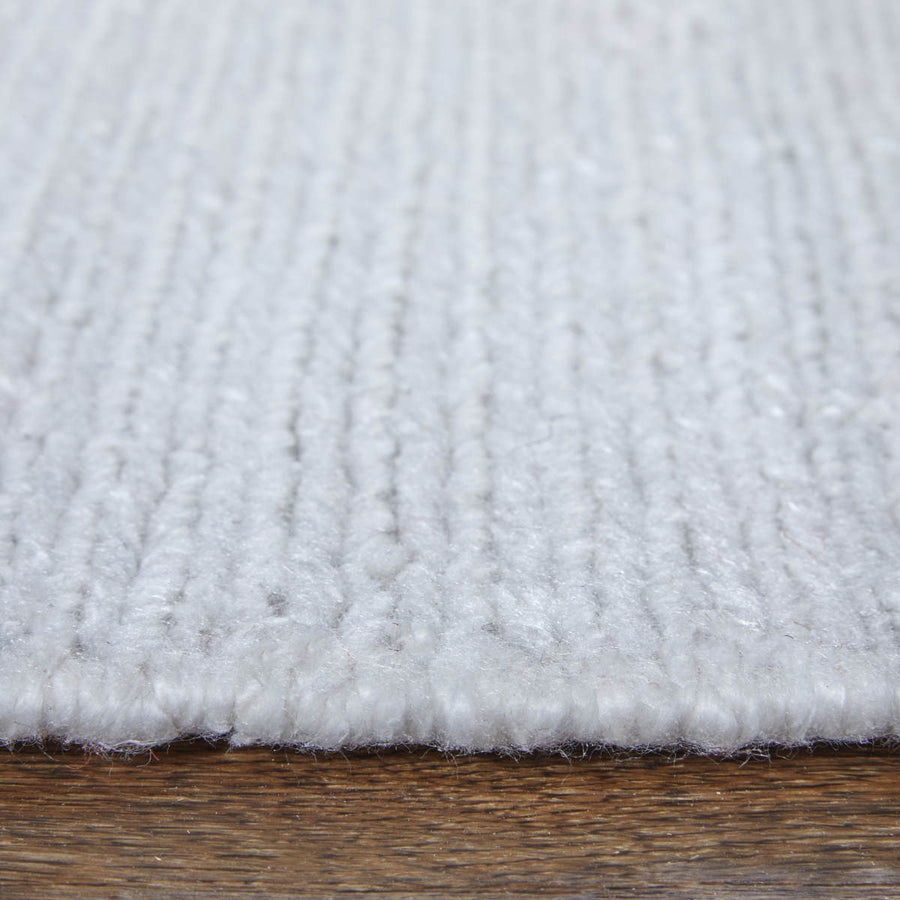 Close-up view of a white, textured area rug with visible woven fibers placed on a wooden floor. The image focuses on the rug’s edge and surface details.