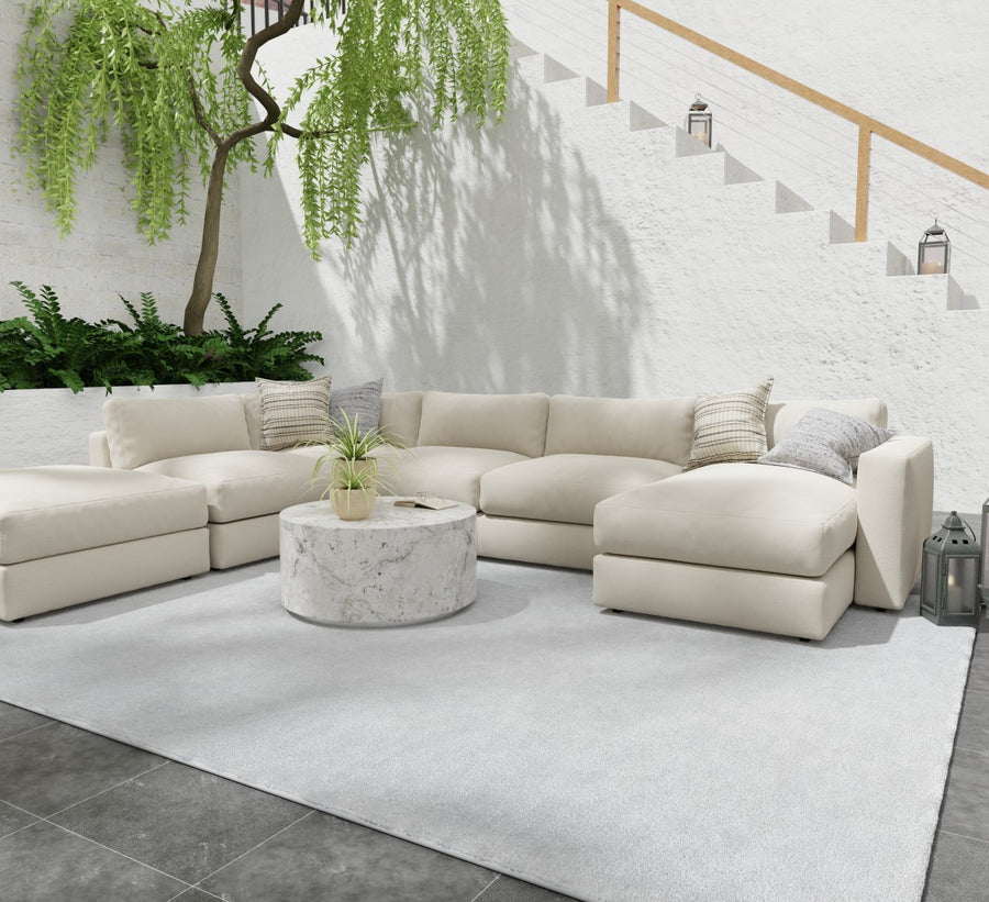 A modern outdoor patio with a large cream sectional sofa, a round marble coffee table, and a light rug. Plants and greenery decorate the area, and white stairs with lanterns ascend in the background.