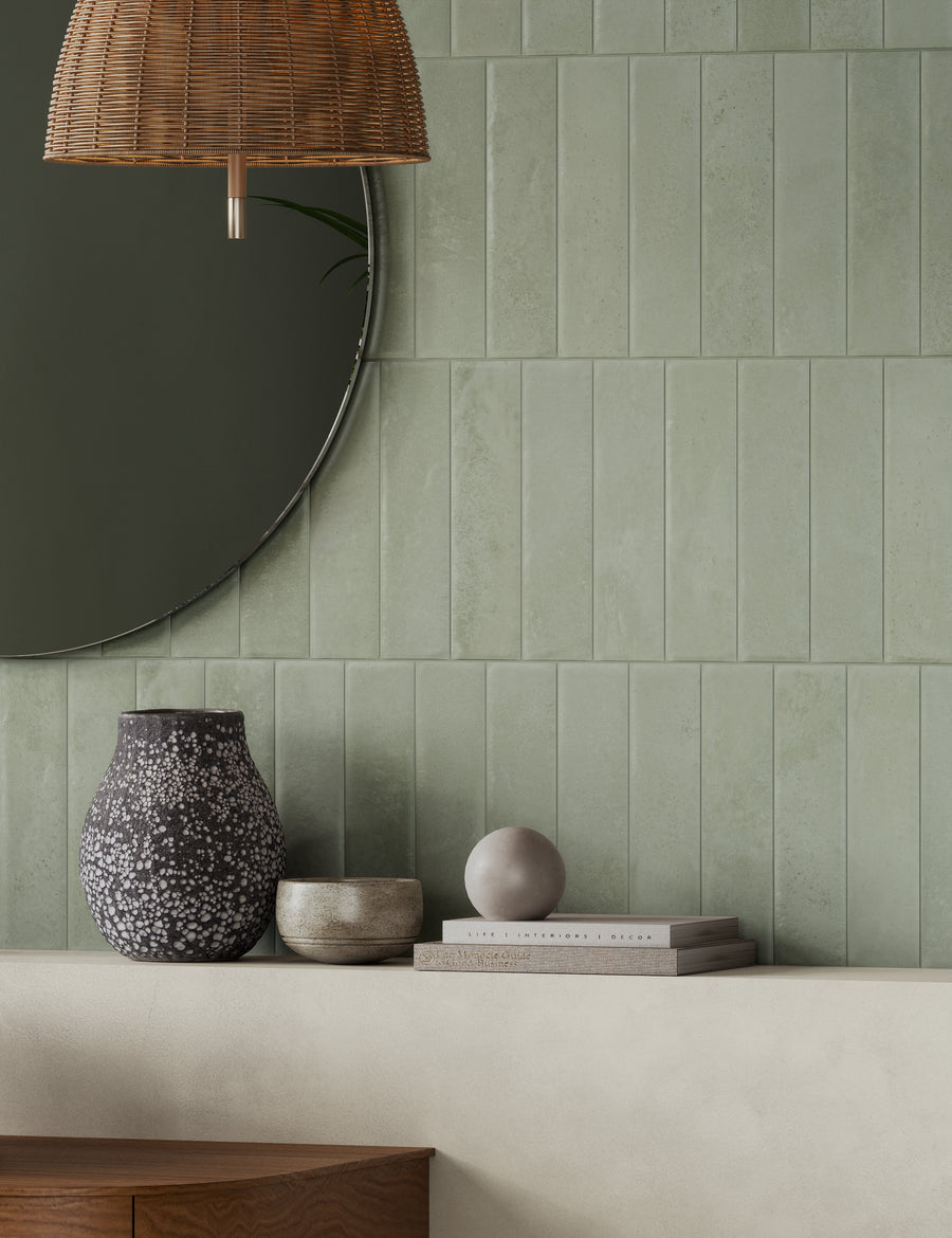 A minimalist interior features a green tiled wall, a round mirror, a woven pendant lamp, and a light wood shelf holding a speckled vase, ceramic bowls, stacked books, and a small gray sphere.