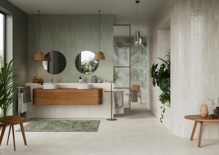 Modern bathroom with green and white tiles, two round mirrors above a wooden double sink vanity, hanging wicker lamps, a green rug, plants, a wooden chair, and a table with decorative items.