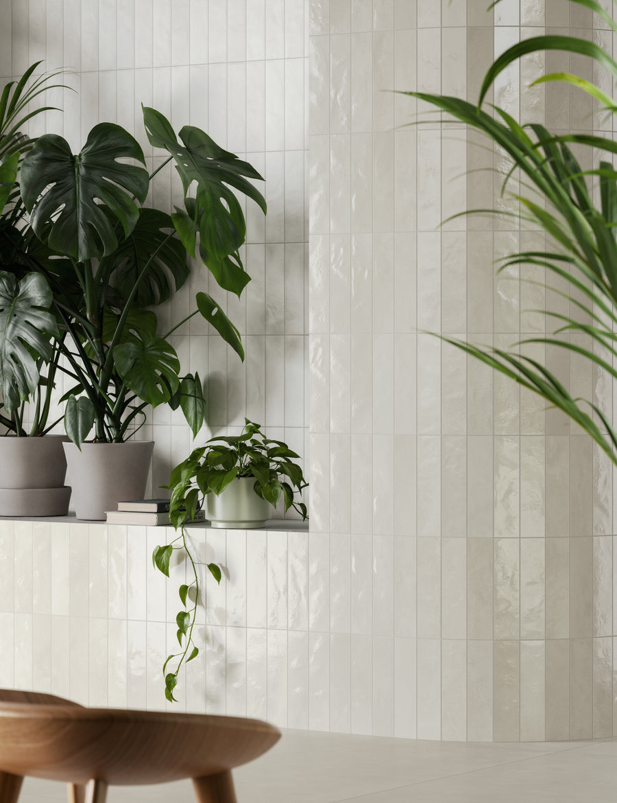Several potted green plants sit on a shelf against a glossy white tiled wall. A wooden stool is partially visible in the foreground, and leafy greenery adds a fresh, modern touch to the minimal space.