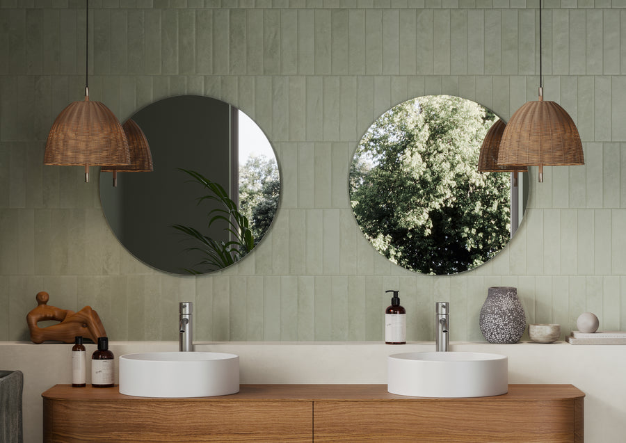 A modern bathroom with two round mirrors above white sinks on a wooden vanity, green tiled walls, wicker pendant lights, and decorative items including soap, a vase, and a wooden sculpture. Trees are reflected in the mirrors.