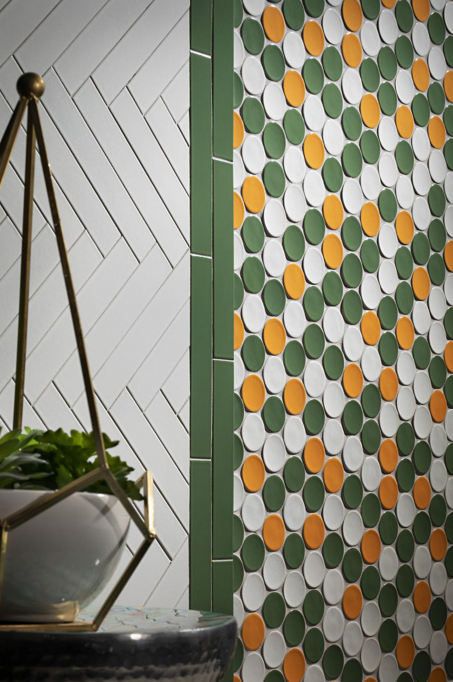 A wall with green vertical tiles and two tile patterns: white chevron on the left and a polka dot design with green, orange, and white circles on the right. A geometric plant holder is in the foreground.