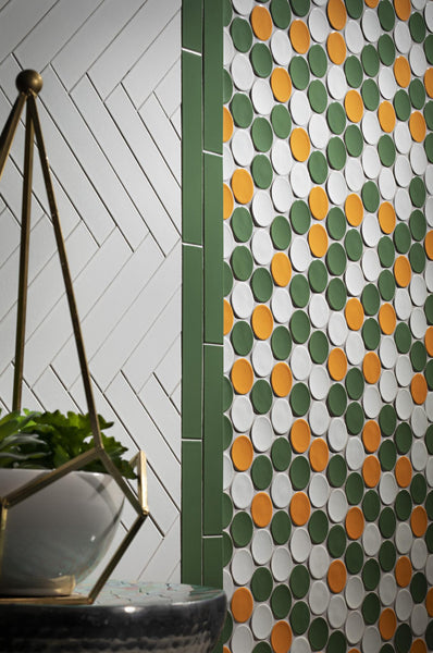 A wall with green vertical tiles and two tile patterns: white chevron on the left and a polka dot design with green, orange, and white circles on the right. A geometric plant holder is in the foreground.