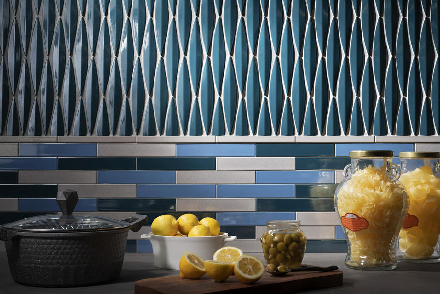 Modern kitchen countertop with a pot, bowl of lemons, a cutting board with sliced lemons and olives, and two glass jars of pasta; blue geometric and brick-patterned tile backsplash.