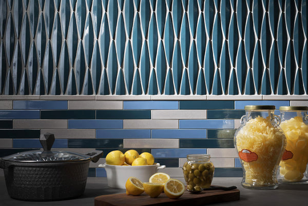 Modern kitchen countertop with a pot, bowl of lemons, a cutting board with sliced lemons and olives, and two glass jars of pasta; blue geometric and brick-patterned tile backsplash.