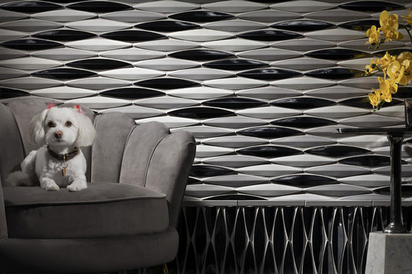 A small white dog with pink bows sits on a gray armchair in front of a modern black, white, and silver geometric tile wall. A vase with yellow flowers is on the right.