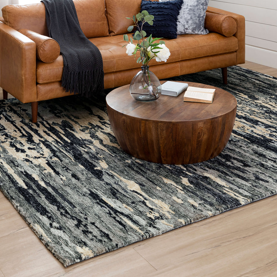 A modern living room with a tan leather sofa, dark accents, a round wood coffee table topped with flowers and a book, and the Bowen Huron Area Rug—a durable polyester rug perfect for high-traffic areas—on light wood floors.