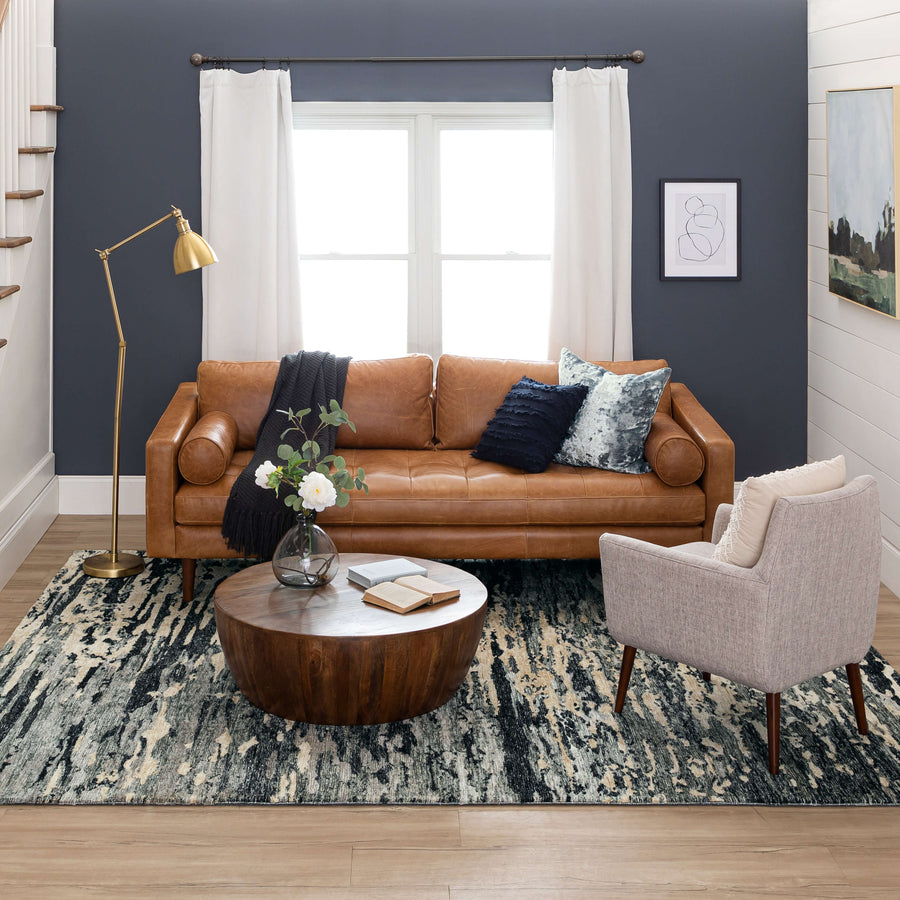 A modern living room with a brown leather sofa, gray armchair, round wooden coffee table with a vase of flowers, floor lamp, abstract rug, dark accent wall, window with white curtains, and framed art on the walls.
