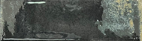 A rectangular Kinetic 2.5x9 Porcelain Subway Tile in the Fluid Force color features a dark, weathered metal appearance with a rough, uneven texture, lighter gray accents, and small spots of rust-like detailing that evoke the aged and distressed look often seen in artisanal pottery or well-worn porcelain tiles.