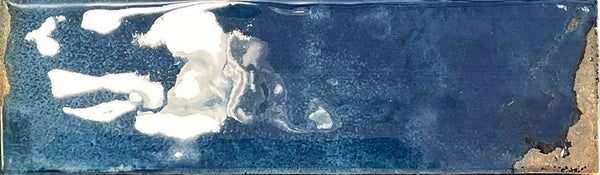 A rectangular Kinetic 2.5x9 Porcelain Subway Tile in the color Rushing Riptides, featuring a blue glossy, reflective glaze reminiscent of artisanal pottery, with lighter and darker blue areas and subtle white reflections on its glazed surface.