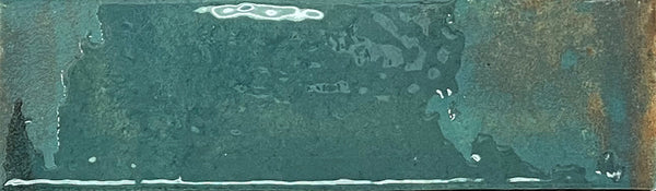 A rectangular Kinetic 2.5x9 Porcelain Subway Tile in the Shifting Skies color, featuring a mottled teal surface with textured variations and patches of brownish rust along the right edge, reminiscent of artisanal pottery finishes.