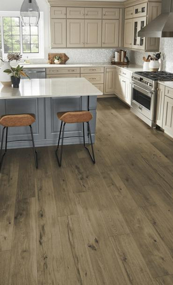 A modern kitchen with light wood cabinets, a large white island with two brown cushioned bar stools, stainless steel appliances, and wide plank wood flooring. Natural light streams in from a window.