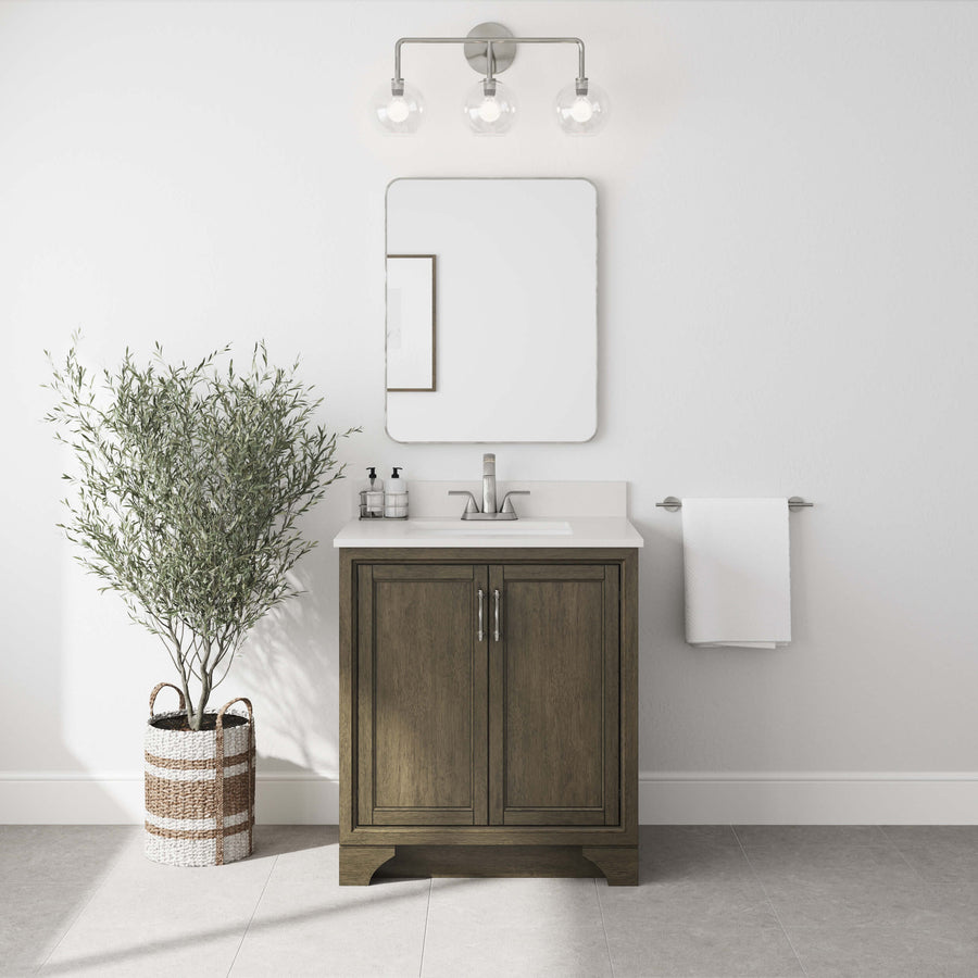 Modern bathroom with a Lanagan 30" Vanity, a rectangular mirror, and a three-bulb light fixture above. A potted plant in a woven basket sits to the left, with a white towel hanging on a rack to the right.