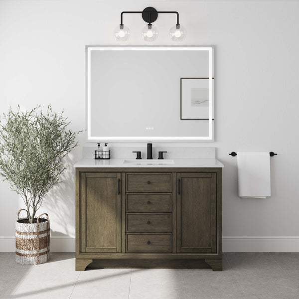 A bathroom with a wooden vanity, a rectangular LED mirror, black faucet, and three-light fixture. A potted plant is on the left, and a towel hangs on the right wall next to framed art.