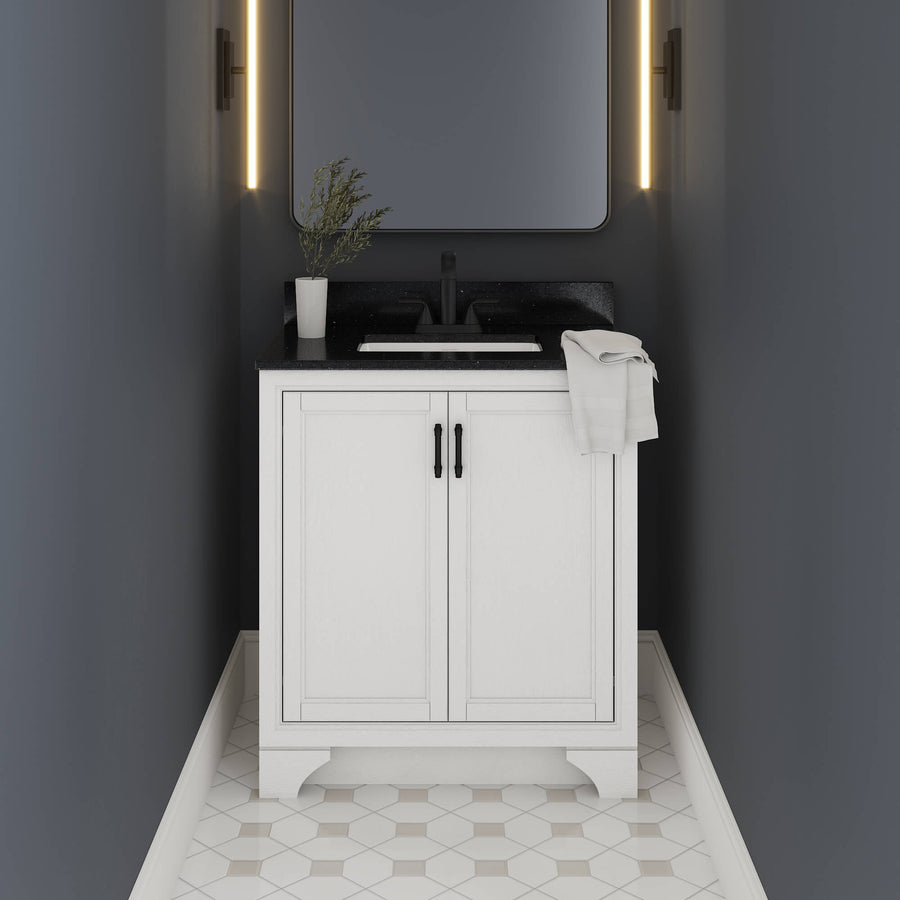 A small modern bathroom with dark walls, a white vanity cabinet, black countertop, rectangular sink, wall-mounted mirror with vertical lights, and geometric tile flooring. A towel and small plant sit on the counter.