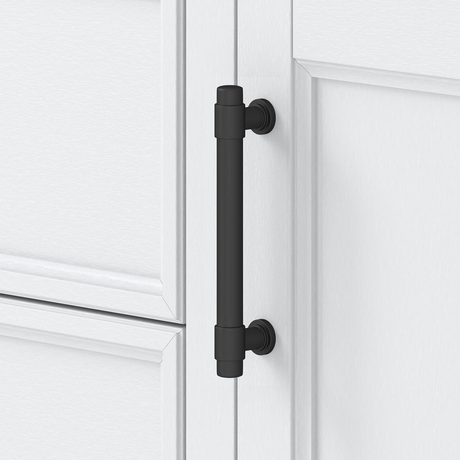 A matte black, cylindrical cabinet handle is mounted vertically on a white cabinet door with raised panel detailing. The handle has round bases at each end and a simple, modern design.