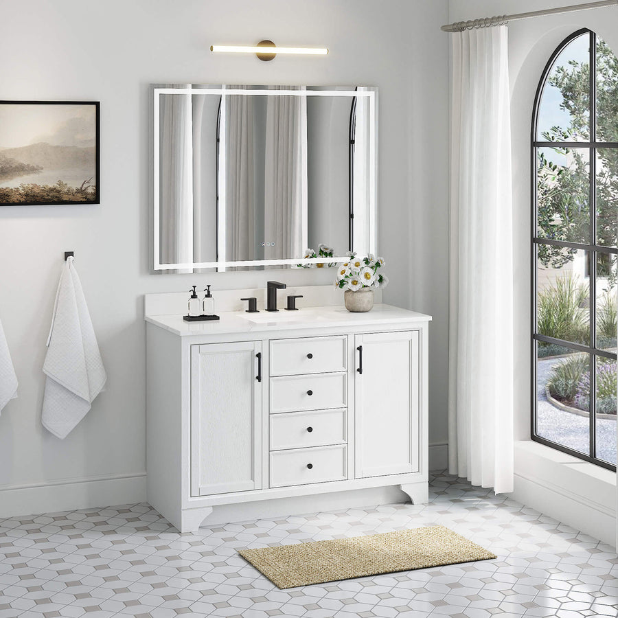 Modern bathroom with a white vanity, dual faucets, and a large mirror. Two white towels hang on the wall, and sunlight streams through a tall arched window with sheer curtains. A beige rug is on the hexagon tile floor.