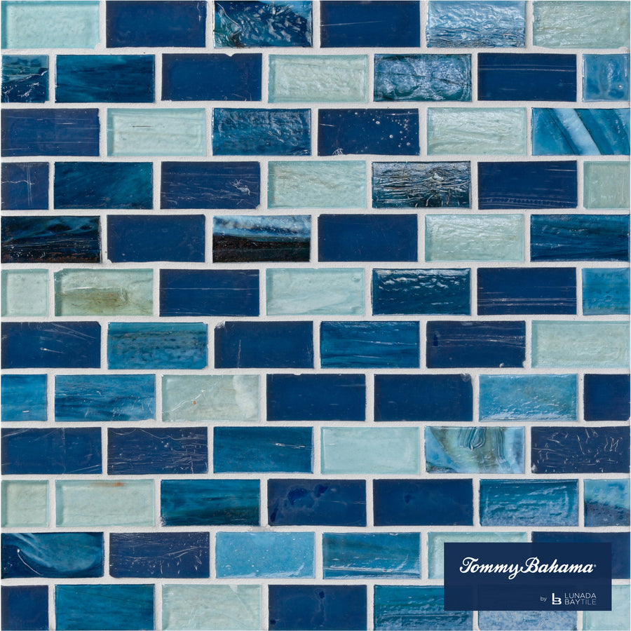 A close-up of a wall with Tommy Bahama 1 sf/sh Brick Ceramic Tile in Lucky Bay Mix, showing rectangular tiles in varying blue and aqua shades. The bottom right has the "Tommy Bahama by Island Style" logo.