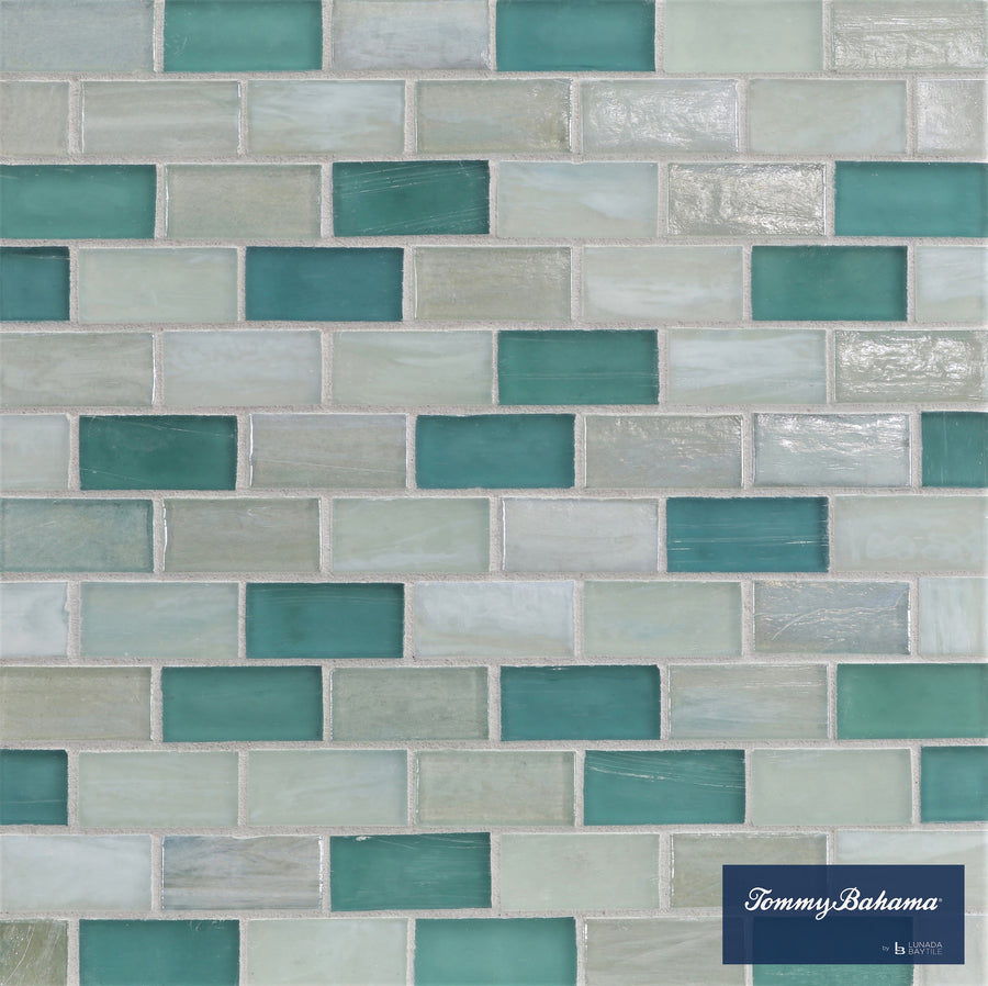 The Tommy Bahama 1 sf/sh Brick Ceramic Tile in San Blas Mix features a mosaic of rectangular tiles in teal, aqua, and off-white shades, arranged in a staggered brick pattern with a small Tommy Bahama logo in the lower right corner.