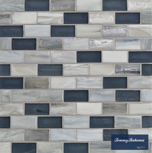 A close-up of a wall with Tommy Bahama 1 sf/sh Brick Ceramic Tile in Twillingate Mix, showing rectangular tiles in blue, gray, and white arranged in a brick pattern. The Tommy Bahama logo appears in the lower right corner.