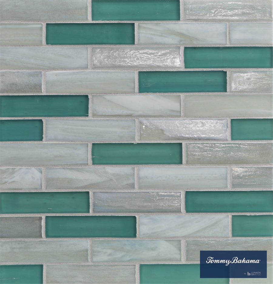 A wall of Tommy Bahama 0.85 sf/sh Brick Ceramic Tile in San Blas Mix—rectangular tiles with aqua, teal, and light gray tones arranged in a brick pattern. A blue label at the bottom right reads Tommy Bahama.