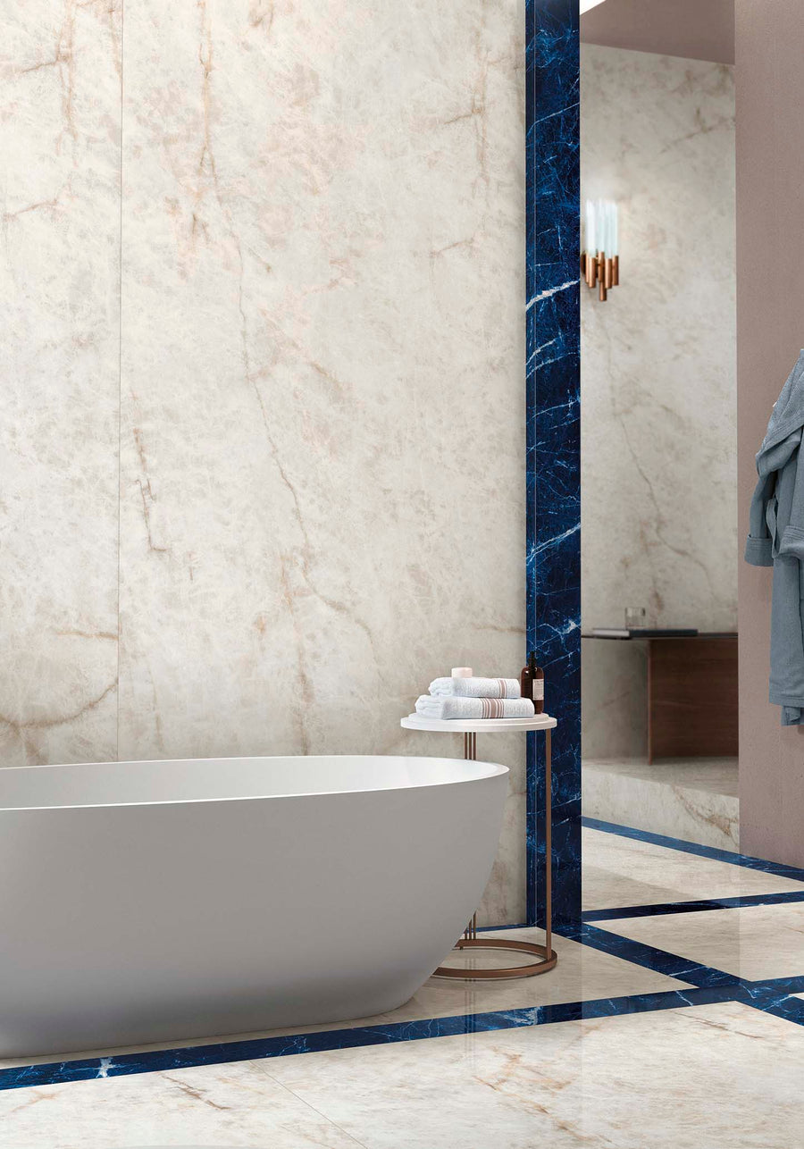 A modern bathroom with a white freestanding bathtub, marble-look walls and floor, blue marble accent trim, a round side table with towels, and a wall-mounted light fixture reflected in the mirror.