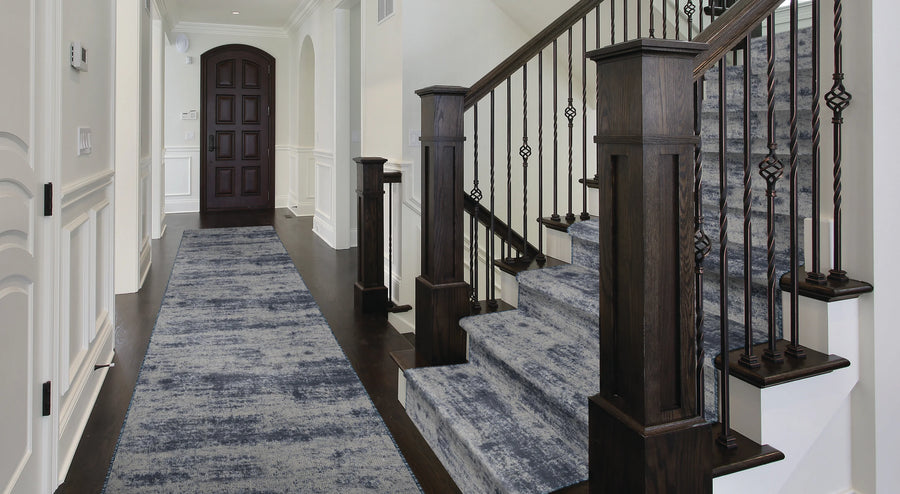 A modern entryway with dark wood floors, a dark wooden staircase with iron railings, and blue-patterned carpet runners on both the hallway and stairs. White walls and trim add brightness to the space.