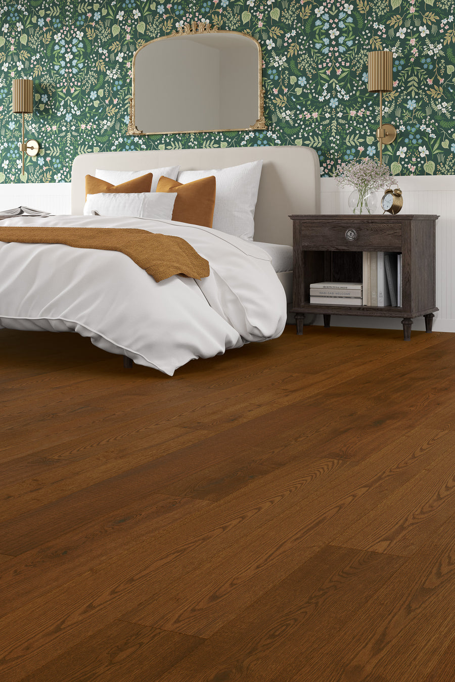 A cozy bedroom with a white bed, gold and white pillows, a dark wooden nightstand, floral green wallpaper, gold wall lamps, a gold-framed mirror, and rich brown hardwood flooring.