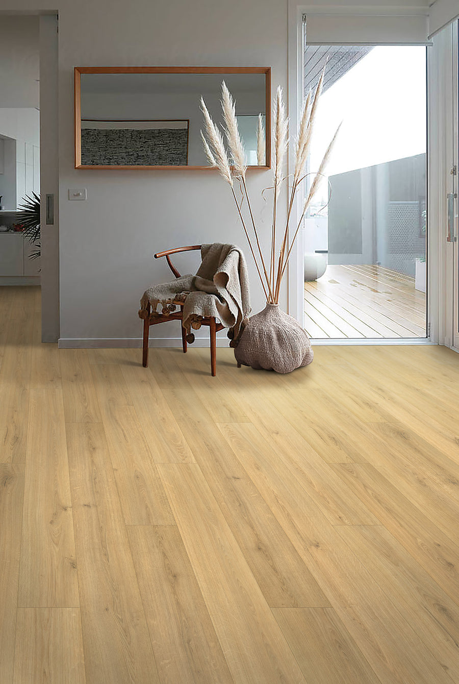 A modern, minimal living space with light wood flooring, a wooden chair draped with a blanket, a large vase holding pampas grass, a wall mirror, and sliding glass doors opening to a balcony.