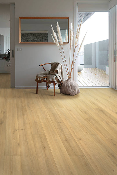 A modern, minimal living space with light wood flooring, a wooden chair draped with a blanket, a large vase holding pampas grass, a wall mirror, and sliding glass doors opening to a balcony.