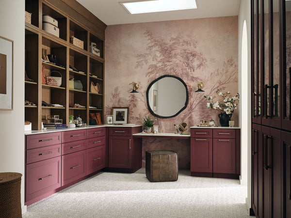 A stylish dressing room with mauve cabinets, open shelves holding shoes and accessories, a round wall mirror, floral wallpaper, decorative items, and a small brown ottoman on a light carpeted floor.