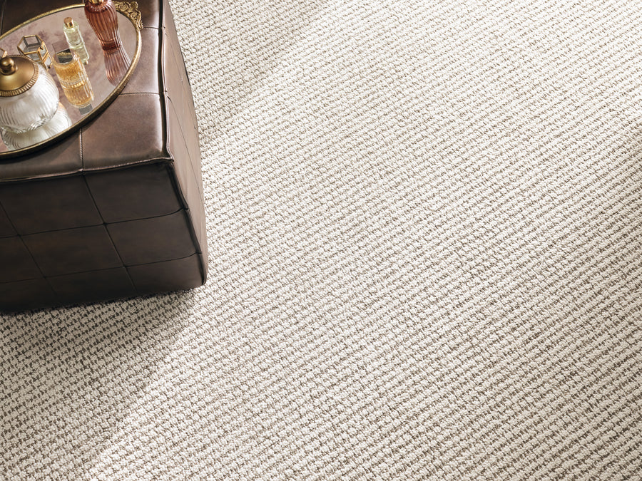 A textured beige carpet covers the floor. In the upper left corner, a brown leather ottoman holds a gold tray with a glass, a bottle, and a small decorative item. Sunlight brightens the scene.