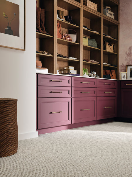 A closet with mauve cabinets and brass handles, open wooden shelves above displaying shoes, bags, and hats, and light carpeted flooring. A framed art print hangs on a white wall to the left.