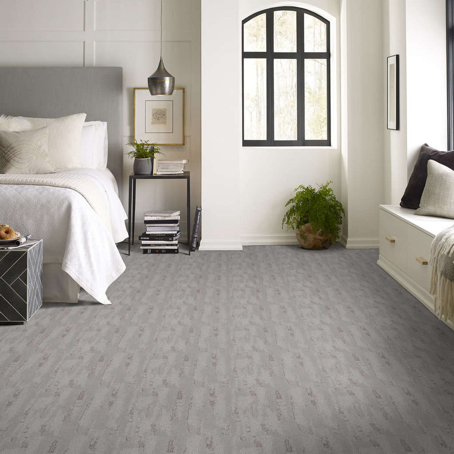 A modern bedroom with a white bed, gray nightstand, potted plants, arched window, pendant light, and light gray textured carpet. Neutral tones create a calm and inviting atmosphere.