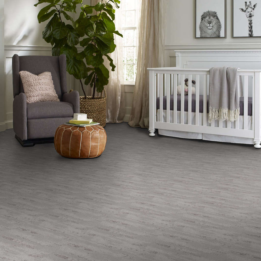 A cozy nursery with a crib, rocking chair with a pillow, leather pouf, large potted plant, and animal-themed artwork on the wall; soft gray flooring and natural light from a window.