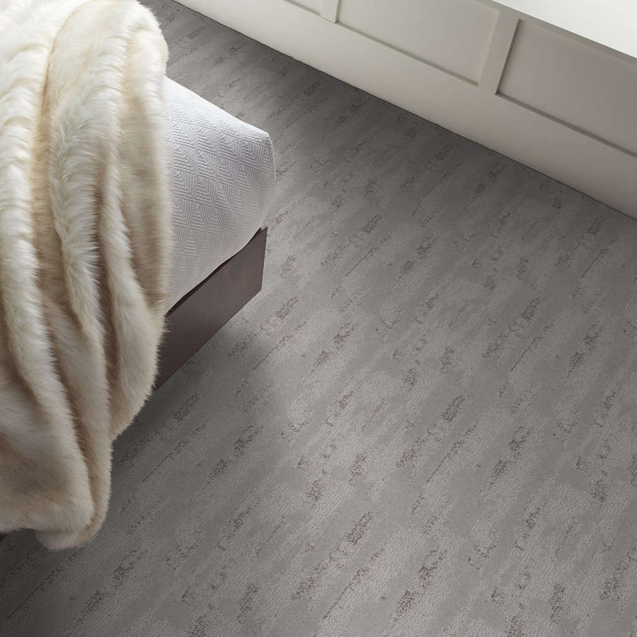 A cozy bedroom corner with a bed covered in a textured white blanket and a soft, light-colored faux fur throw, next to a floor with light grey wood-look flooring.