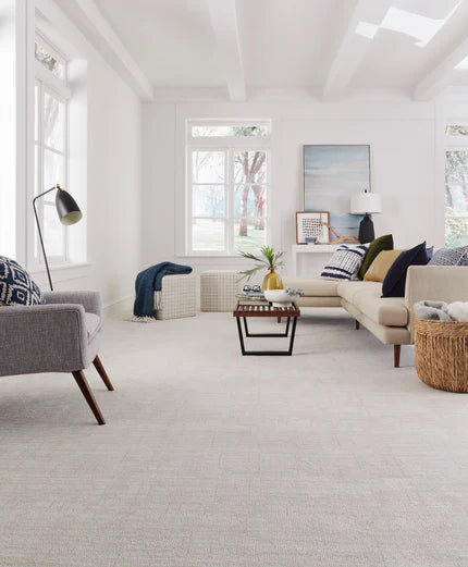 Bright, modern living room with white walls and carpet, a beige sectional sofa with colorful pillows, a gray armchair, a coffee table, and large windows letting in natural light. Decor includes a basket and framed art.