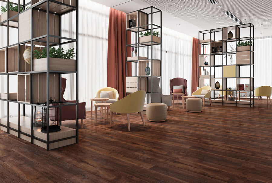 Modern lounge area with wooden floors, open metal shelves containing plants and decor, light-colored chairs and ottomans, small tables, and large windows with sheer white curtains and rust-colored drapes.