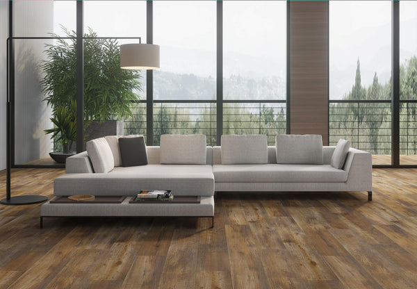 A modern living room with a large gray sectional sofa, wooden floors, a tall potted plant, a floor lamp, and large windows showing trees and mountains outside.