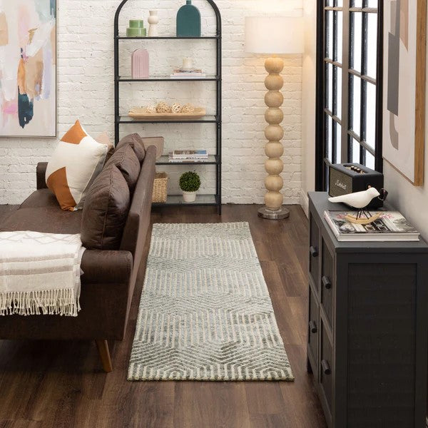 A cozy living room with a brown sofa, patterned rug, gray cabinet, lamp with round wooden base, black shelf with decor, wall art, and large window with grid design letting in natural light.