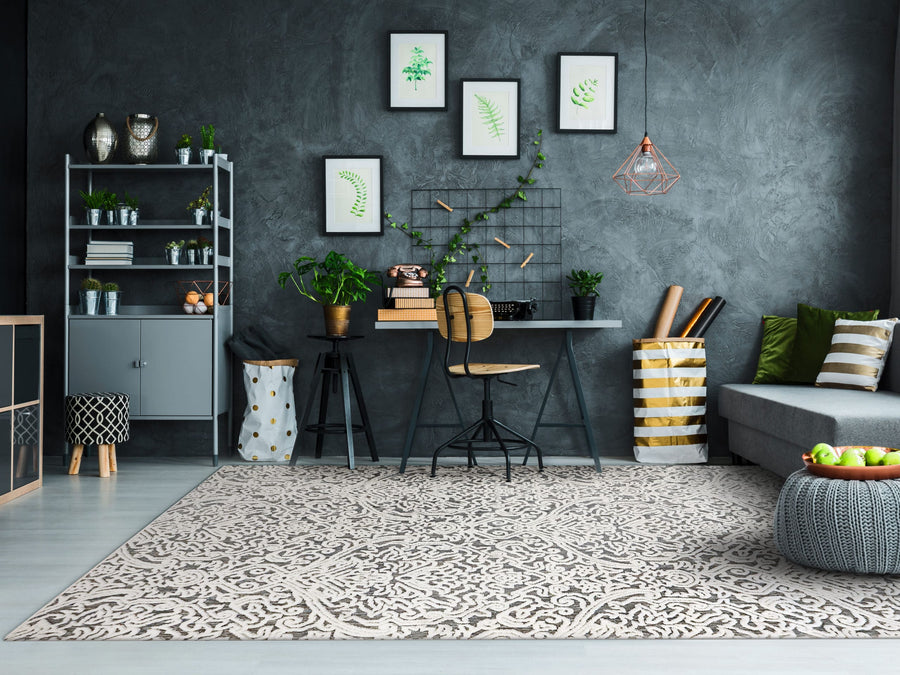 A modern home office with a patterned rug, a desk with a wooden chair, plants, a gray sofa with cushions, and framed botanical prints on a dark gray wall. Shelves and storage baskets decorate the space.