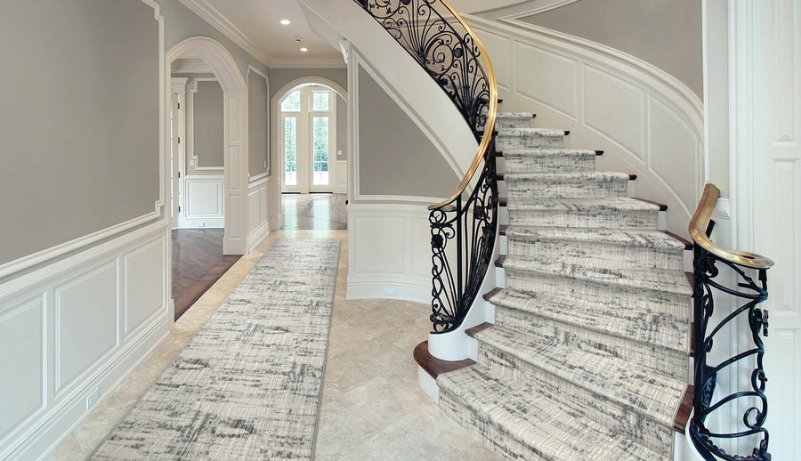 Elegant foyer with a curved staircase featuring ornate black railing and a light-colored, patterned carpet runner. Neutral walls, arched doorway, wainscoting, and natural light from large double doors.