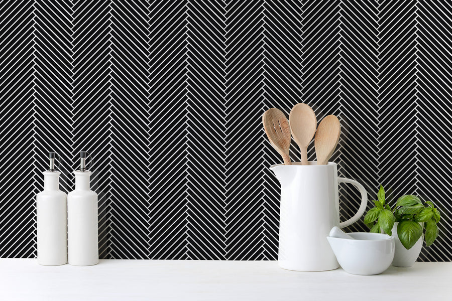 White ceramic kitchenware, wooden utensils, and a small basil plant rest on a white surface before a black wall adorned with Luna Chevron Glass Tile, creating an elegant monochrome look with its striking white chevron pattern.