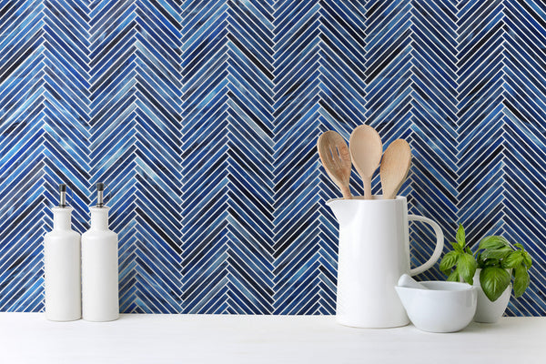 White kitchen utensils in a jug, a small bowl, and a basil plant sit on a white counter in front of the Luna Chevron Glass Tile backsplash, creating a clean monochromatic look with stylish blue glass accents.