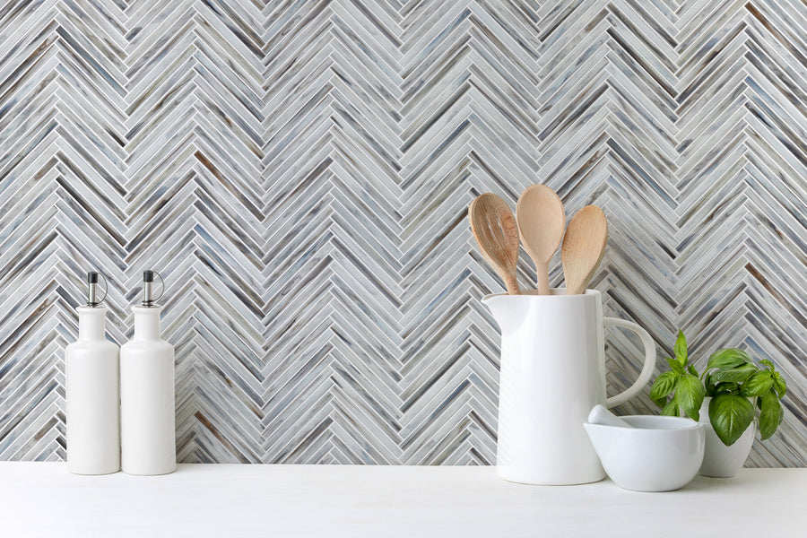 A modern kitchen counter features a Luna Chevron Glass Tile backsplash, a white utensils holder with wooden spoons, two white bottles, a basil plant, and a white mortar and pestle for timeless monochromatic style.